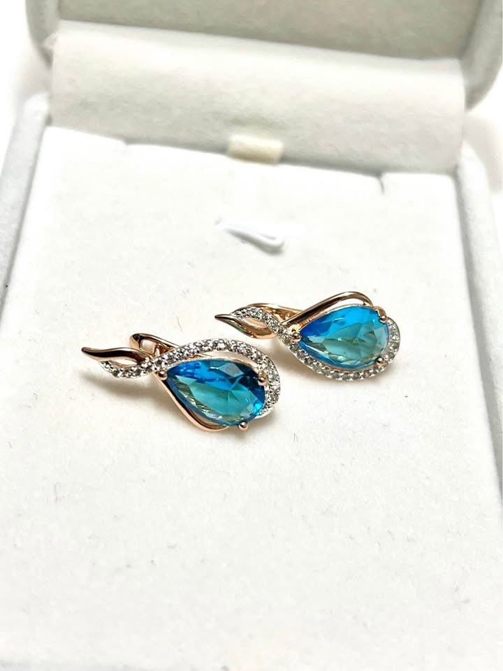 A close-up of 14k rolled rose gold earrings with a faceted blue topaz and a row of crystals.