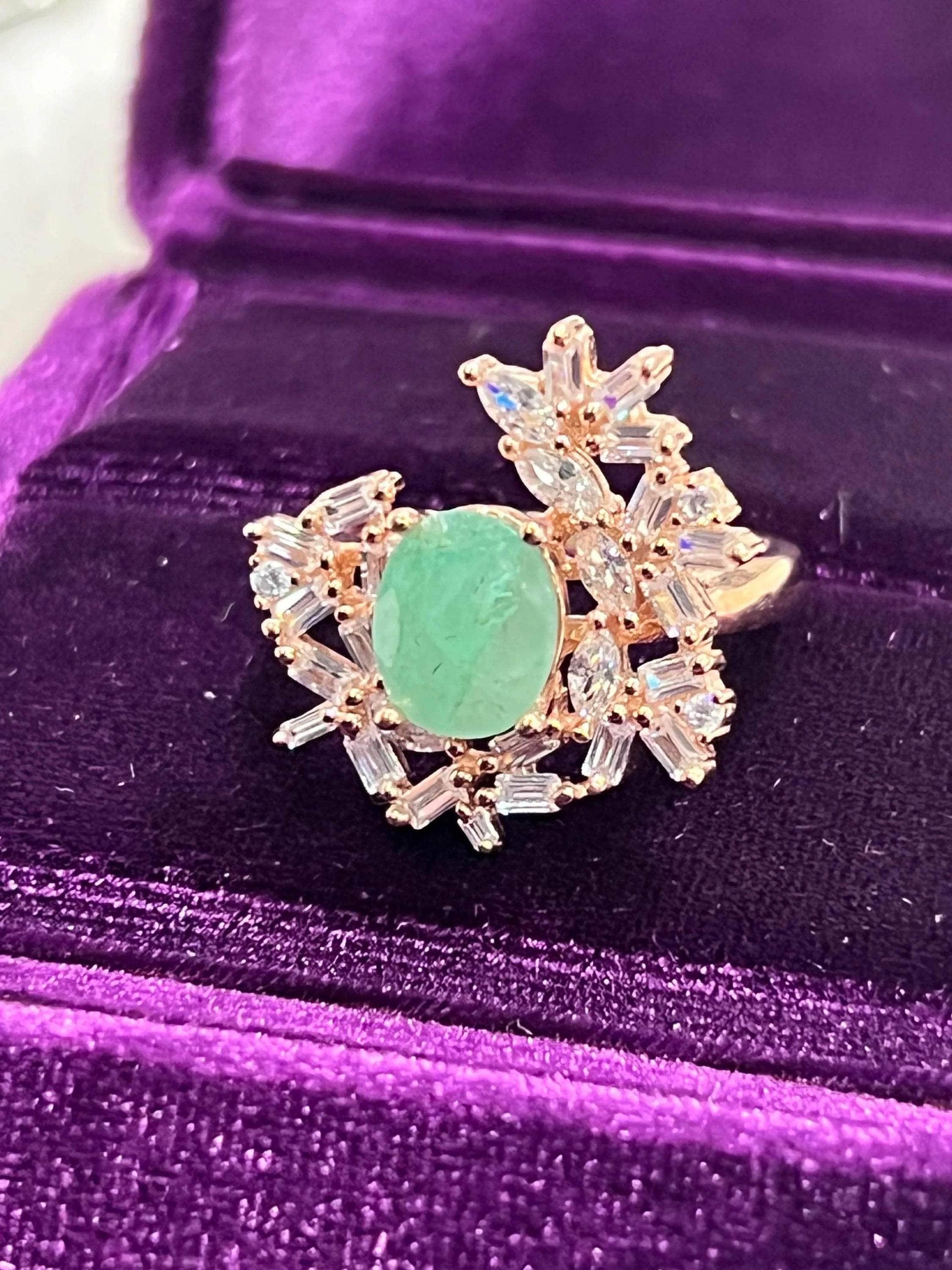Exquisite vintage-style ring with a central emerald gemstone surrounded by sparkling sapphires, in a purple box.