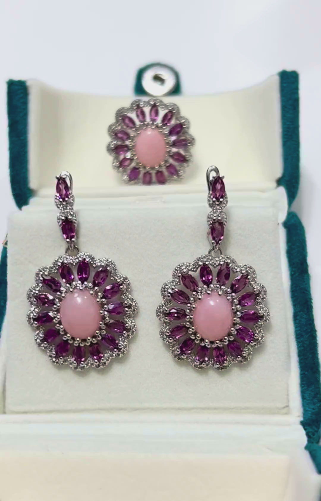 A video showcasing a matching jewelry set featuring a ring and a pair of dangle earrings, each adorned with a central pink opal surrounded by a halo of vibrant Rhodolite garnets and sparkling accent stones, displayed in a white jewelry box.