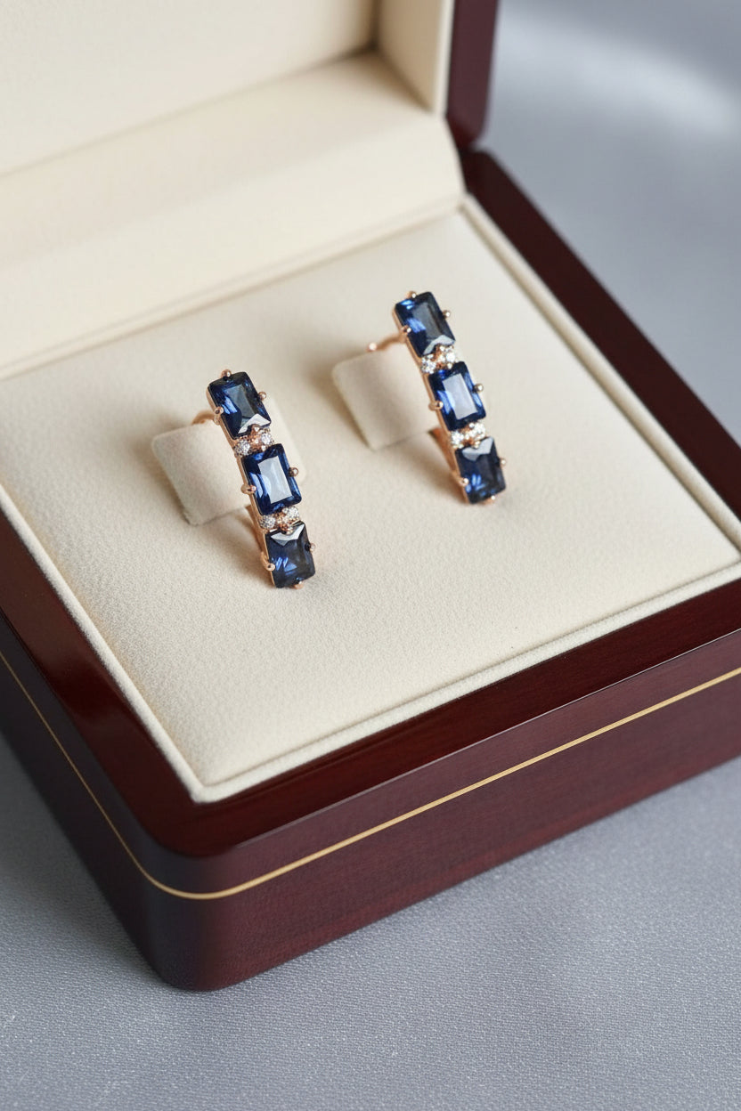 Pair of 14K rose gold-filled earrings with brilliant emerald-cut blue sapphire stones, held by fingers.