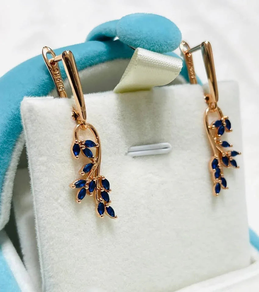 A pair of 14K rose gold stamped earrings with a leafy design and natural blue sapphire stones, on a display.