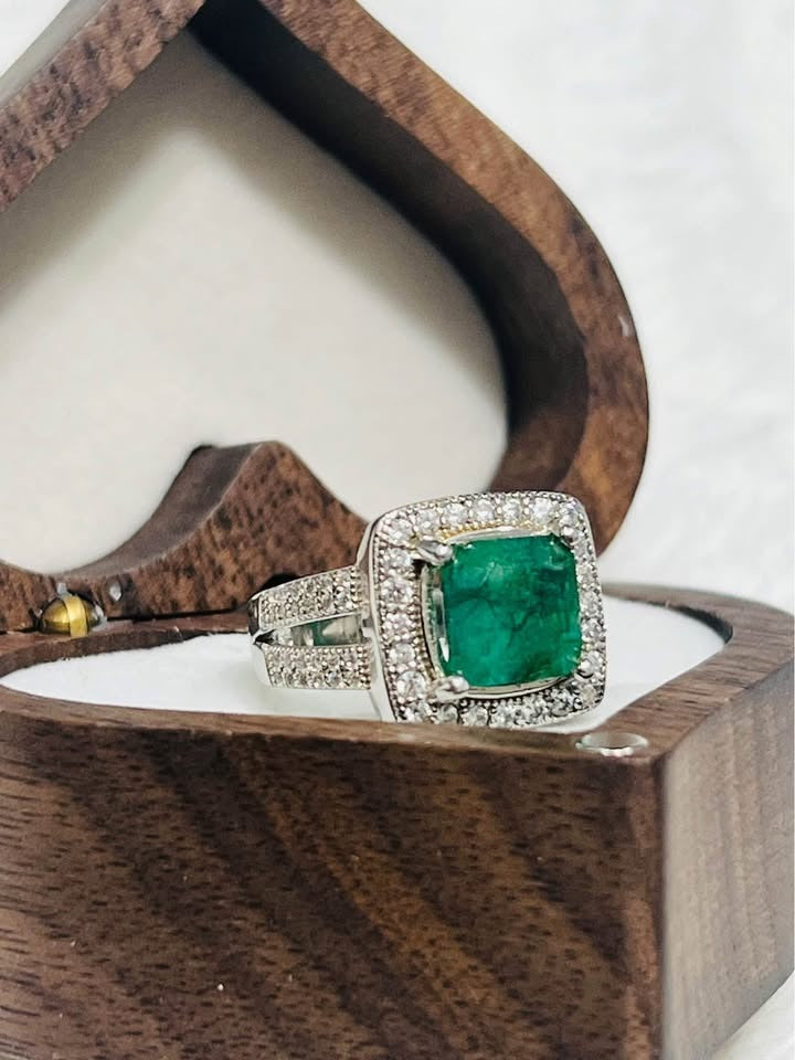 Side angle of a 2 CT natural emerald and diamond halo ring with pave band, resting in a wooden heart box.