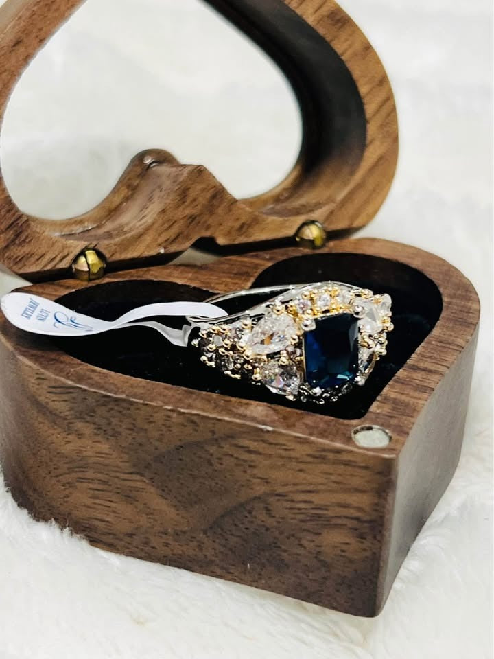 Angled view of a shining sapphire and diamond ring in an open heart-shaped wooden box with tag.