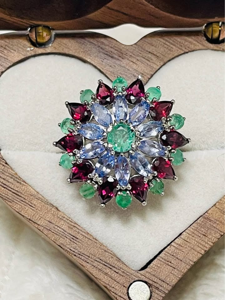 A detailed close-up of a floral ring. The central emerald is surrounded by marquise-cut tanzanite and teardrop garnets.