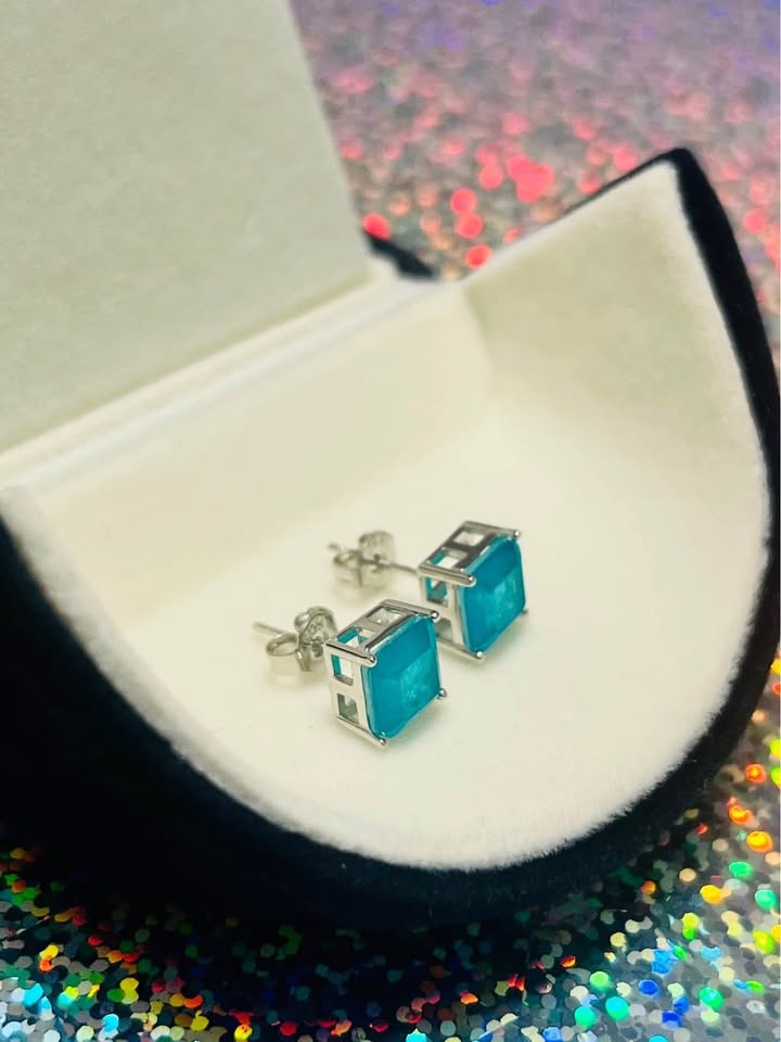 Dainty genuine blue topaz square stud earrings in sterling silver, presented in a black jewelry box on a shimmering surface.