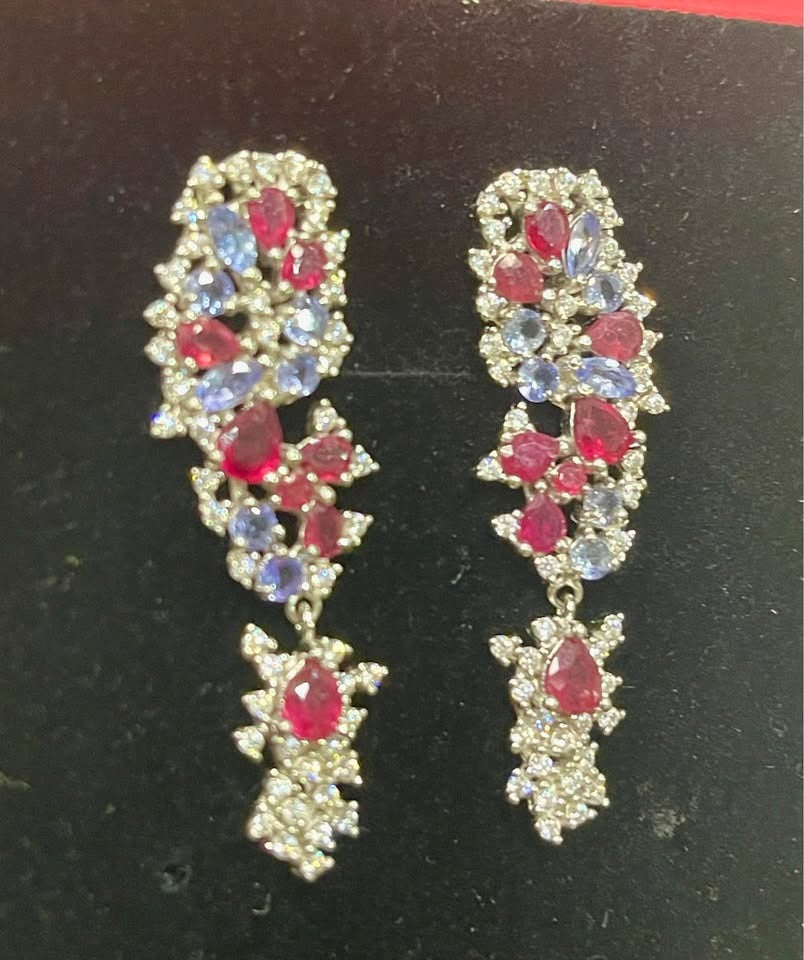 A close-up view of intricate dangle earrings. They are adorned with tanzanite, ruby, and brilliant white gemstones.