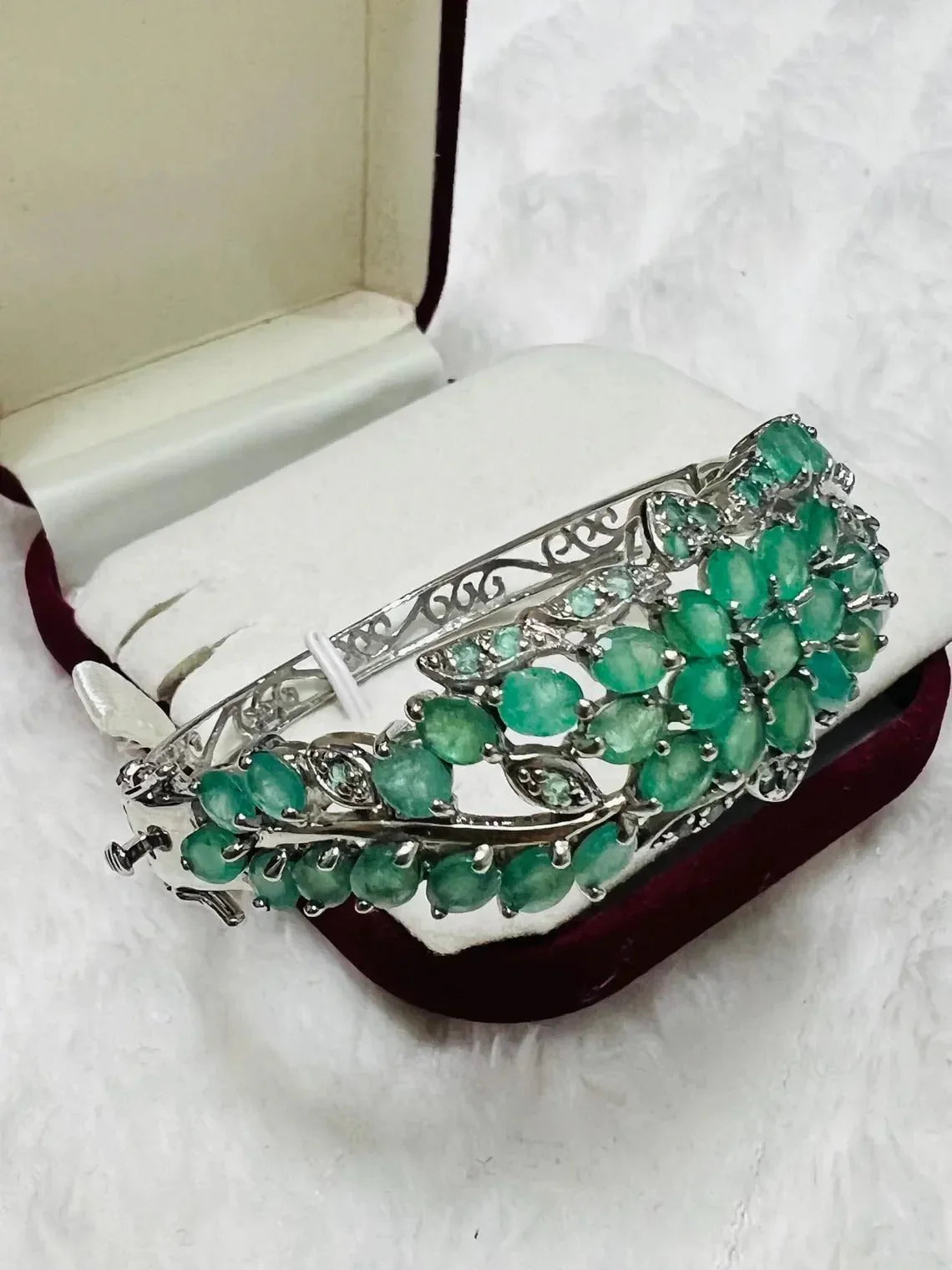 Angled view of a vibrant emerald and tanzanite floral bangle in a red velvet jewelry box, highlighting design.