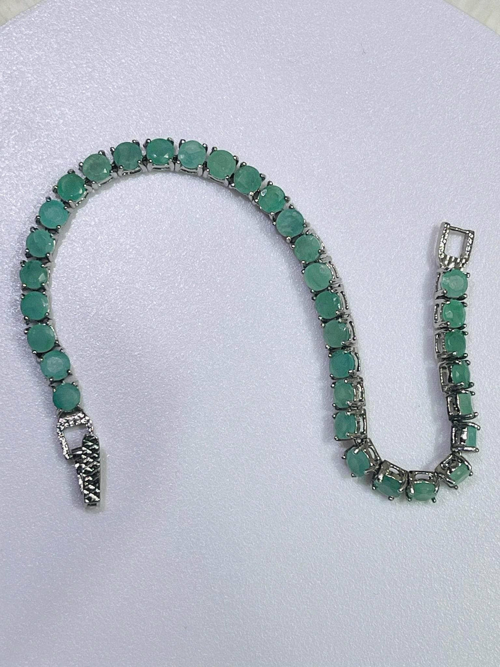 Elegant silver tennis bracelet adorned with numerous round, vibrant emerald gemstones, displayed unclasped on a white surface.