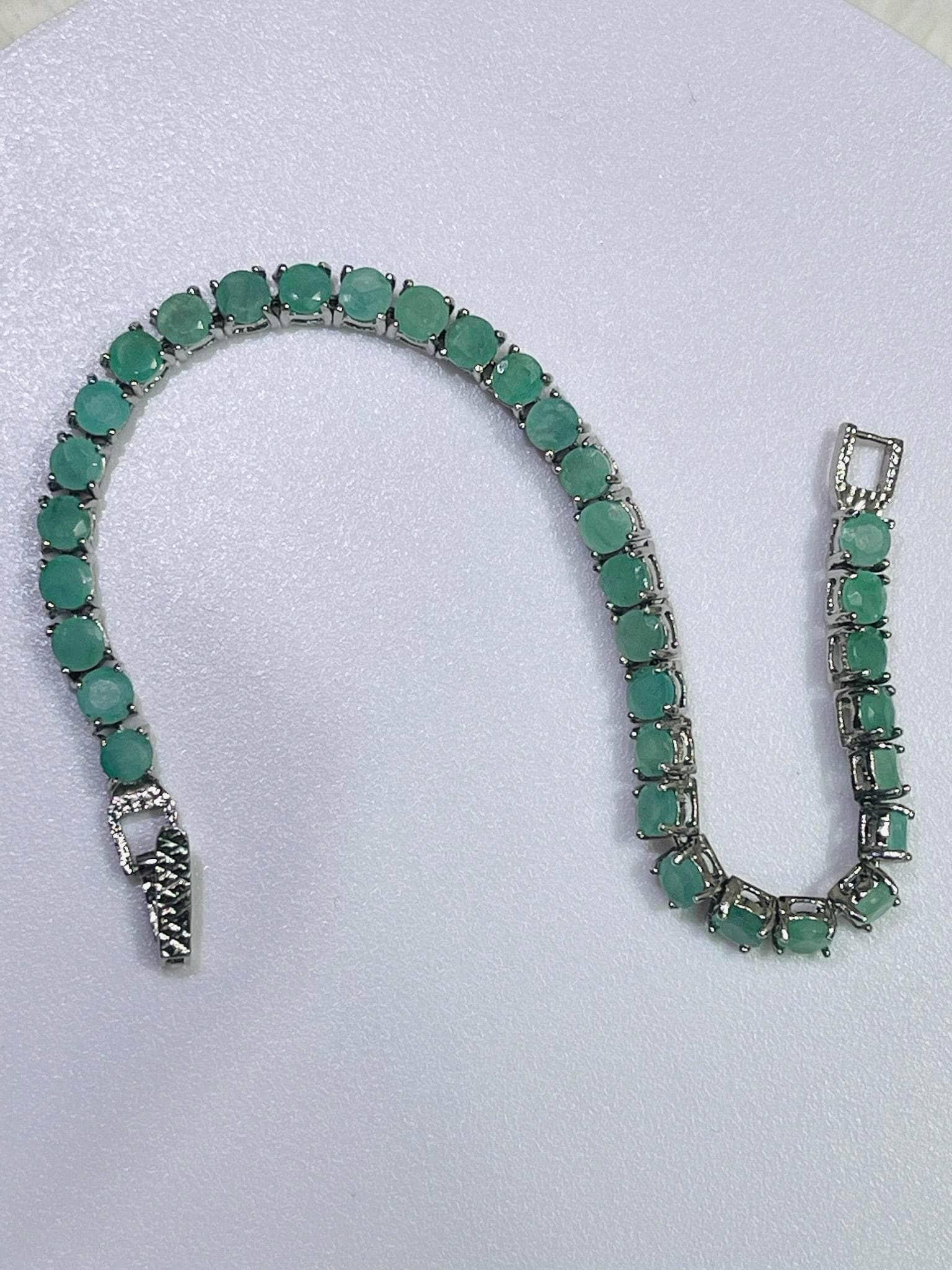 Elegant silver tennis bracelet adorned with numerous round, vibrant emerald gemstones, displayed unclasped on a white surface.