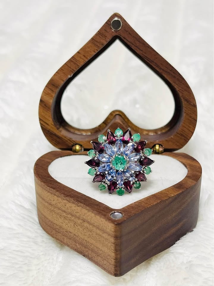 A stunning floral ring in a heart-shaped box. The design features mix of tanzanite, emerald, and rhodolite garnet gemstones.