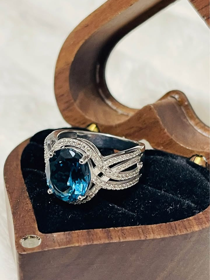 Side angle of an oval London Blue Topaz and diamond ring, resting elegantly in an open wooden box.