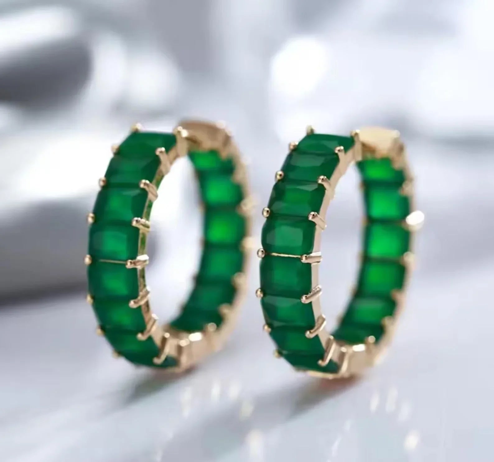 Matching pair of deep green matrix emerald hoop earrings with gold-tone settings on a bright surface.