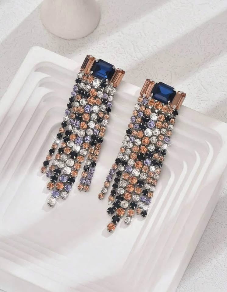 A top-down view of multi-colored waterfall earrings on a white tray. The elegant design features a large blue centerpiece.