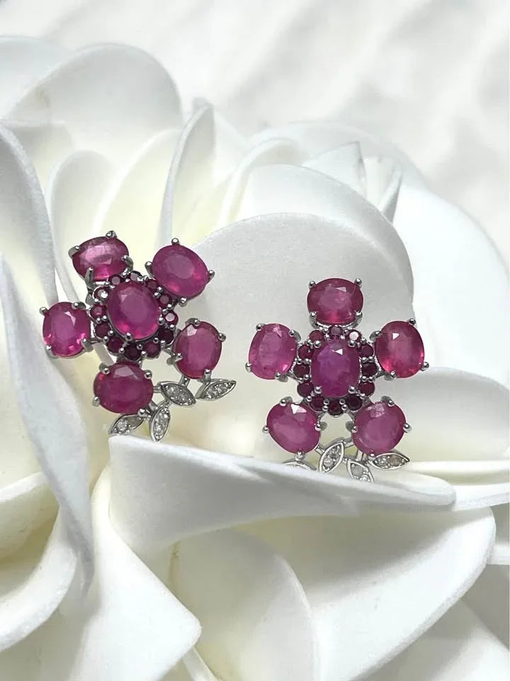 A pair of floral-inspired sterling silver stud earrings featuring natural pink rubies, resting on a white flower.