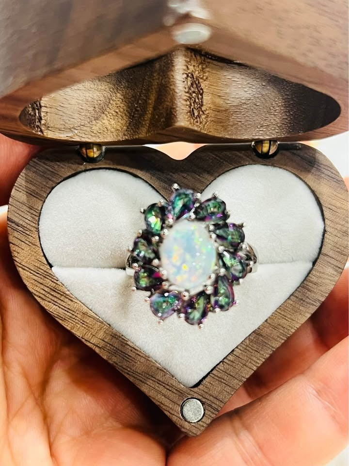 A hand holding an open wooden heart-shaped box, displaying a beautiful opal and mystic topaz ring.