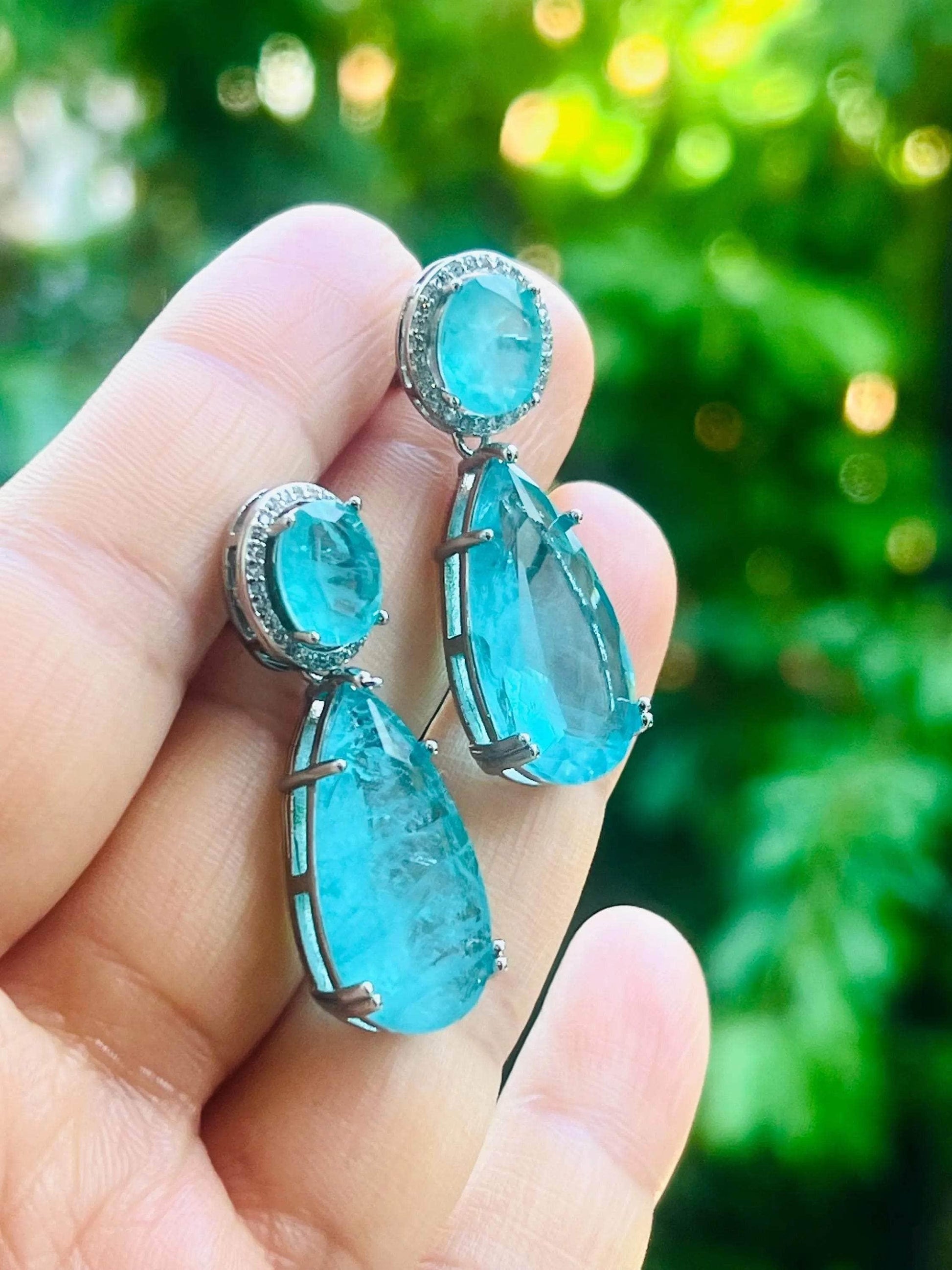 Close-up of Paraiba tourmaline dangle earrings in hand, capturing their vibrant color against nature.