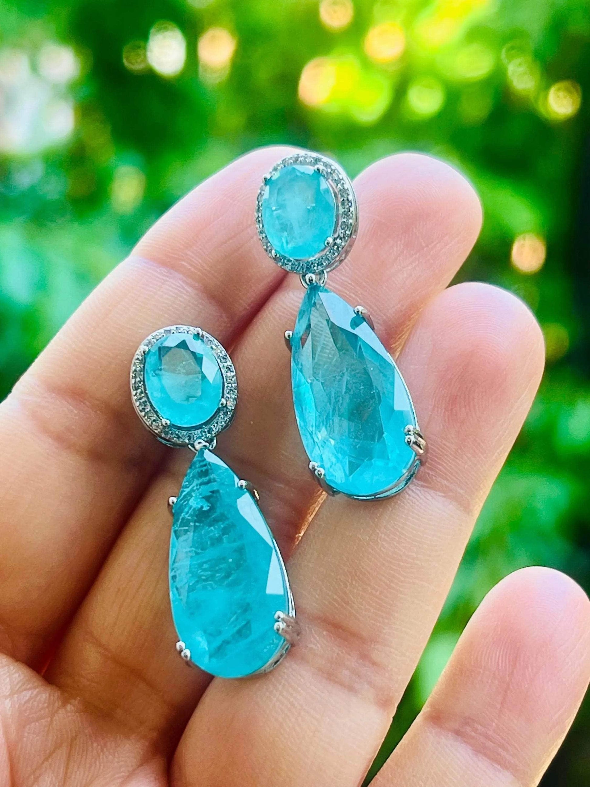 Bright Paraiba tourmaline earrings held in a hand against a blurred green outdoor background, showing vibrancy.