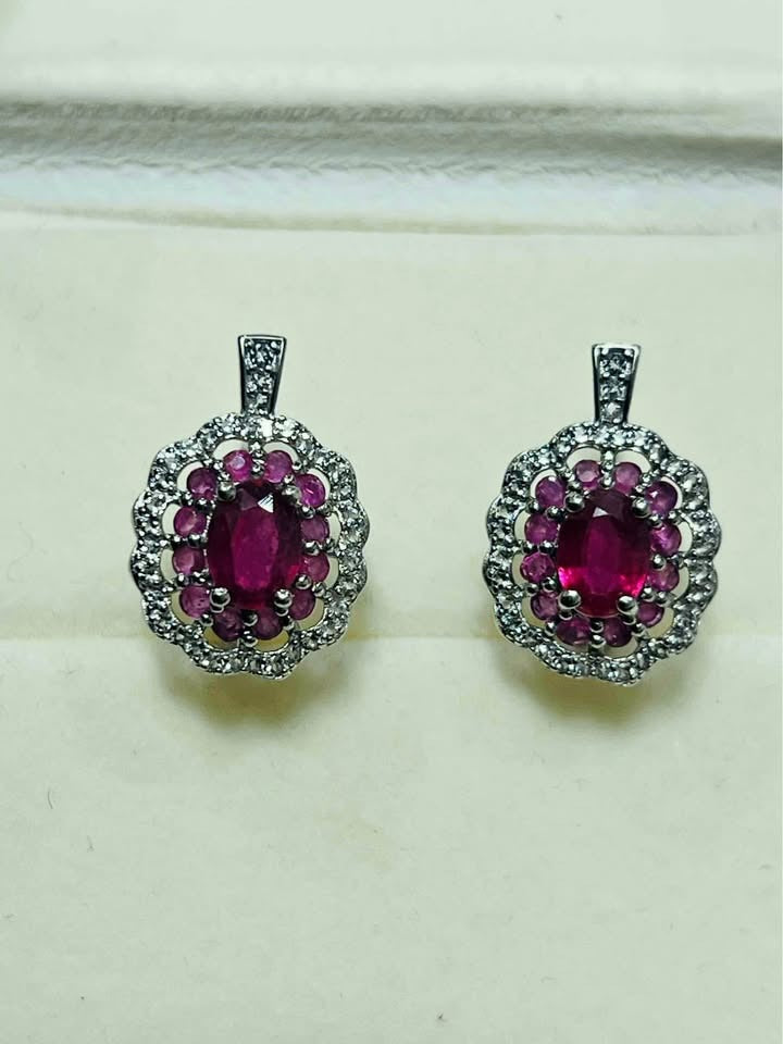 Pair of rhodium-plated sterling silver ruby halo earrings with vibrant oval rubies surrounded by clear stones.