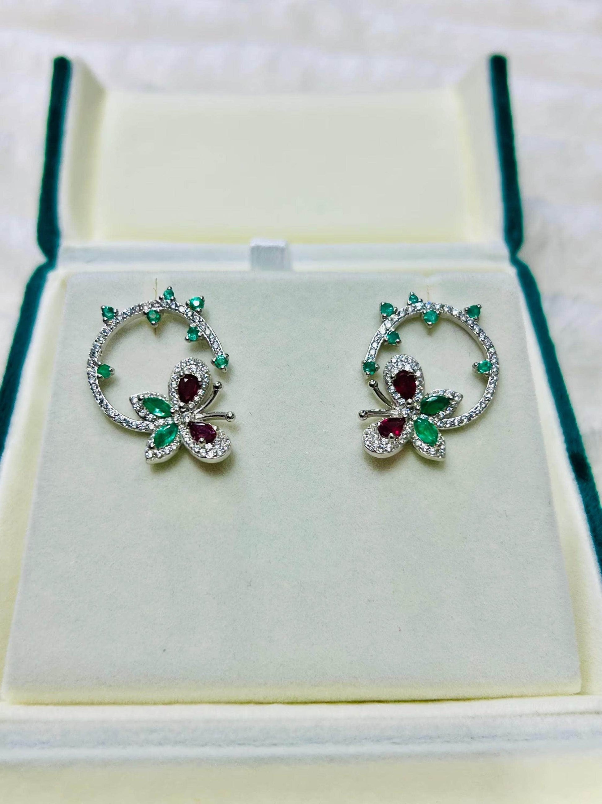 Pair of silver butterfly and flower earrings with emerald and ruby gemstones, displayed in a white jewelry box.