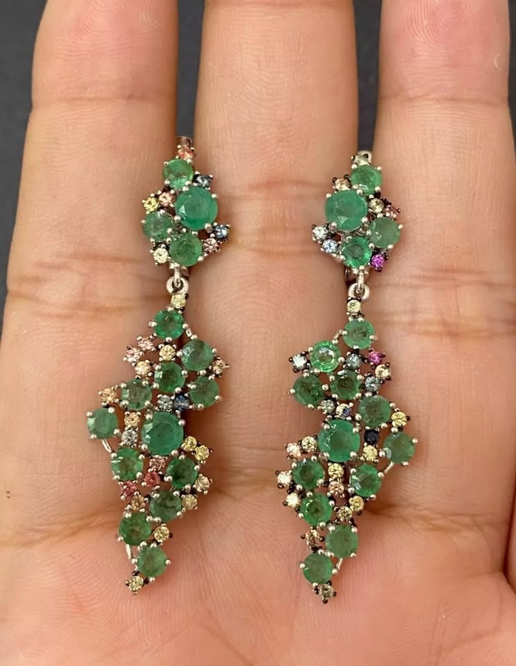 Close-up of a sterling silver drop earring, highlighting the vivid green emeralds and deep blue sapphires.