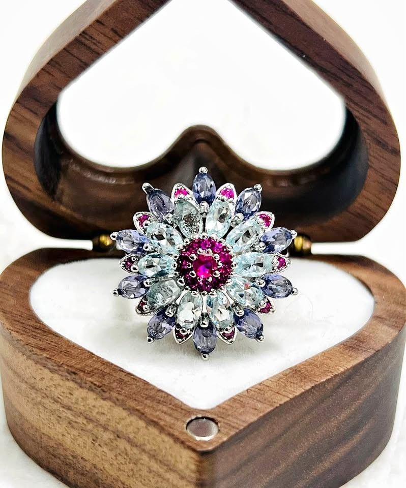 Sterling silver floral ring with blue and purple gemstones and a pink center, displayed in a wooden heart box.