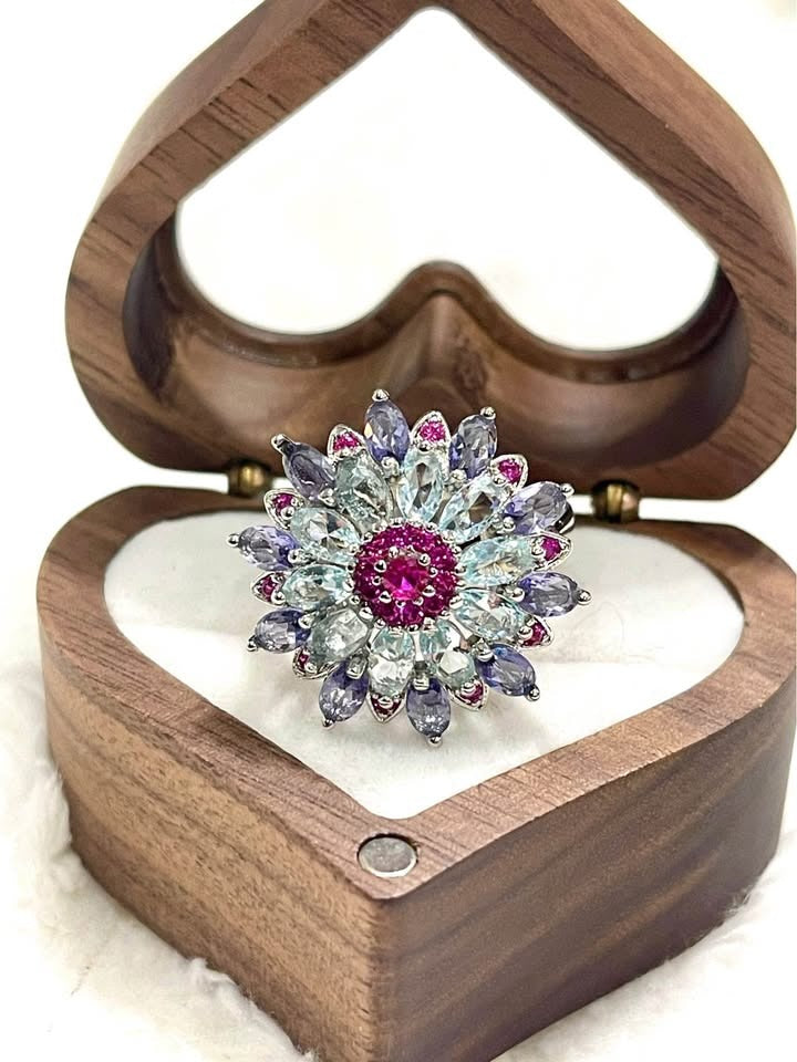  Top-down view of a sterling silver ring with a flower design made of multicolored gemstones in a heart-shaped box.