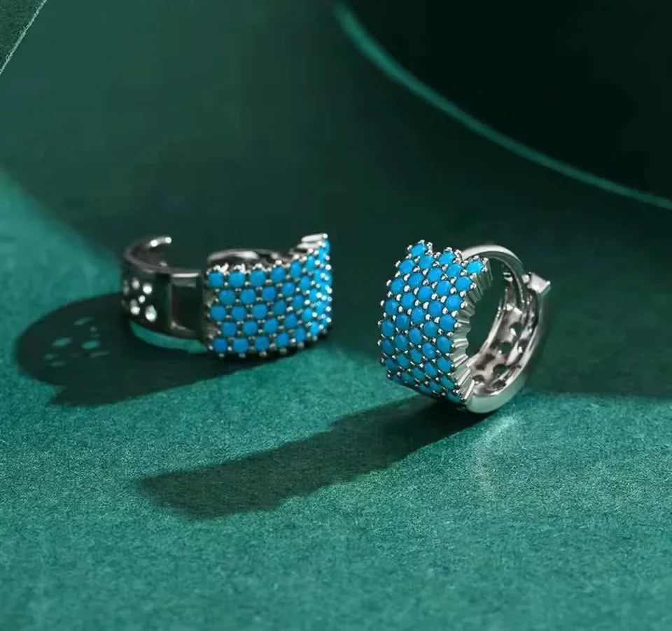 Product shot of two wide sterling silver hoop earrings, featuring real turquoise pave stones on a green background.
