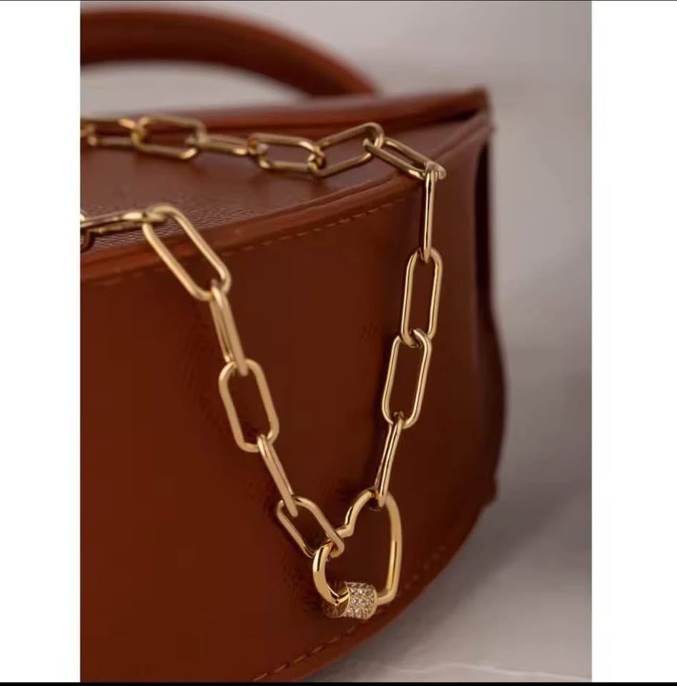 Vermeil paperclip chain necklace with a heart pave clasp draped on a stylish brown leather bag.