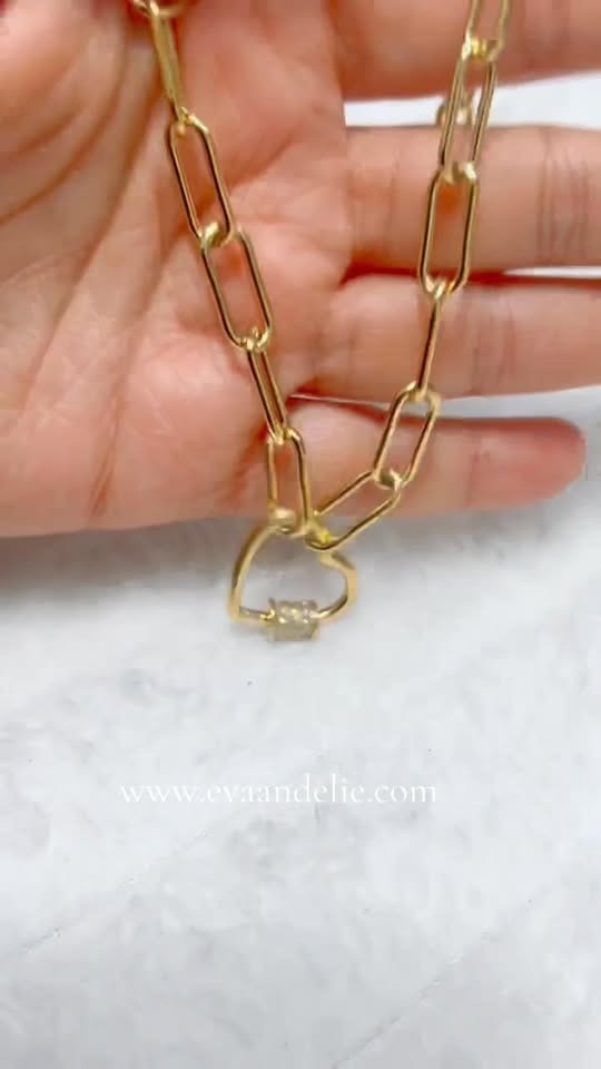 Hand showcasing a vermeil paperclip chain necklace with a heart pave clasp, website visible.