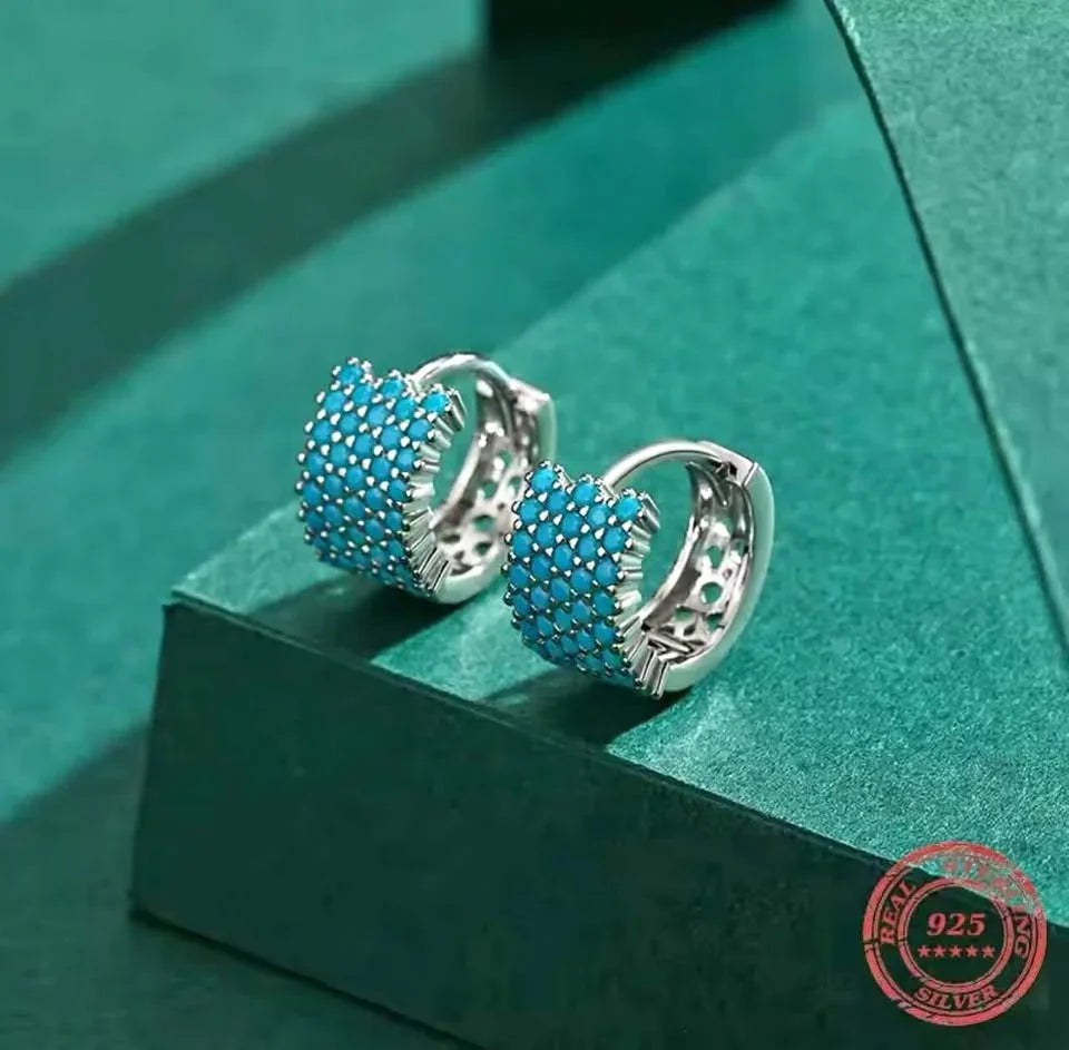 A pair of wide sterling silver hoop earrings with a vibrant pave of real turquoise, displayed on a green surface.