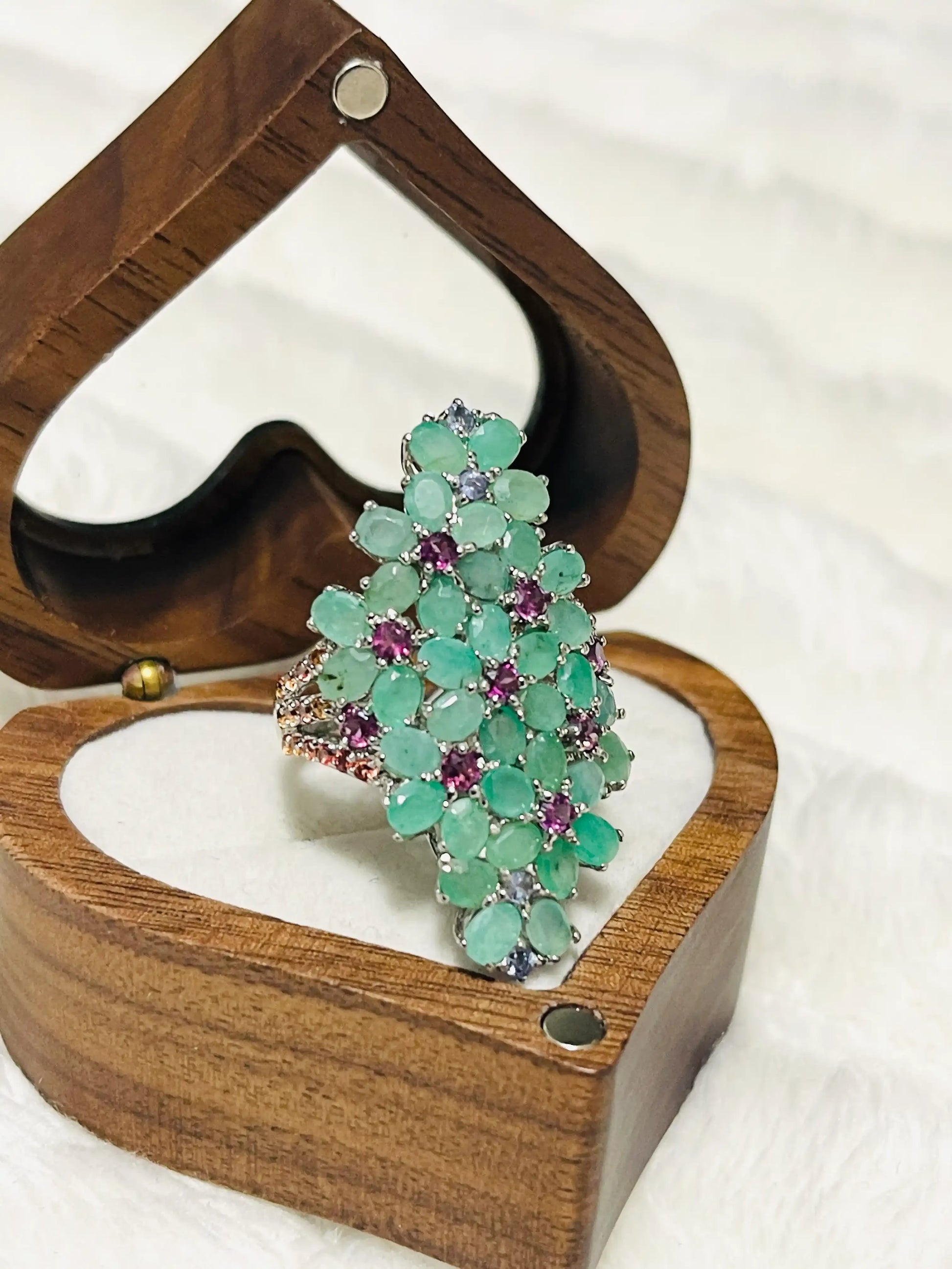 Angled view of a vibrant cluster ring, adorned with green emeralds and purple amethysts, showcased in a wooden heart box.