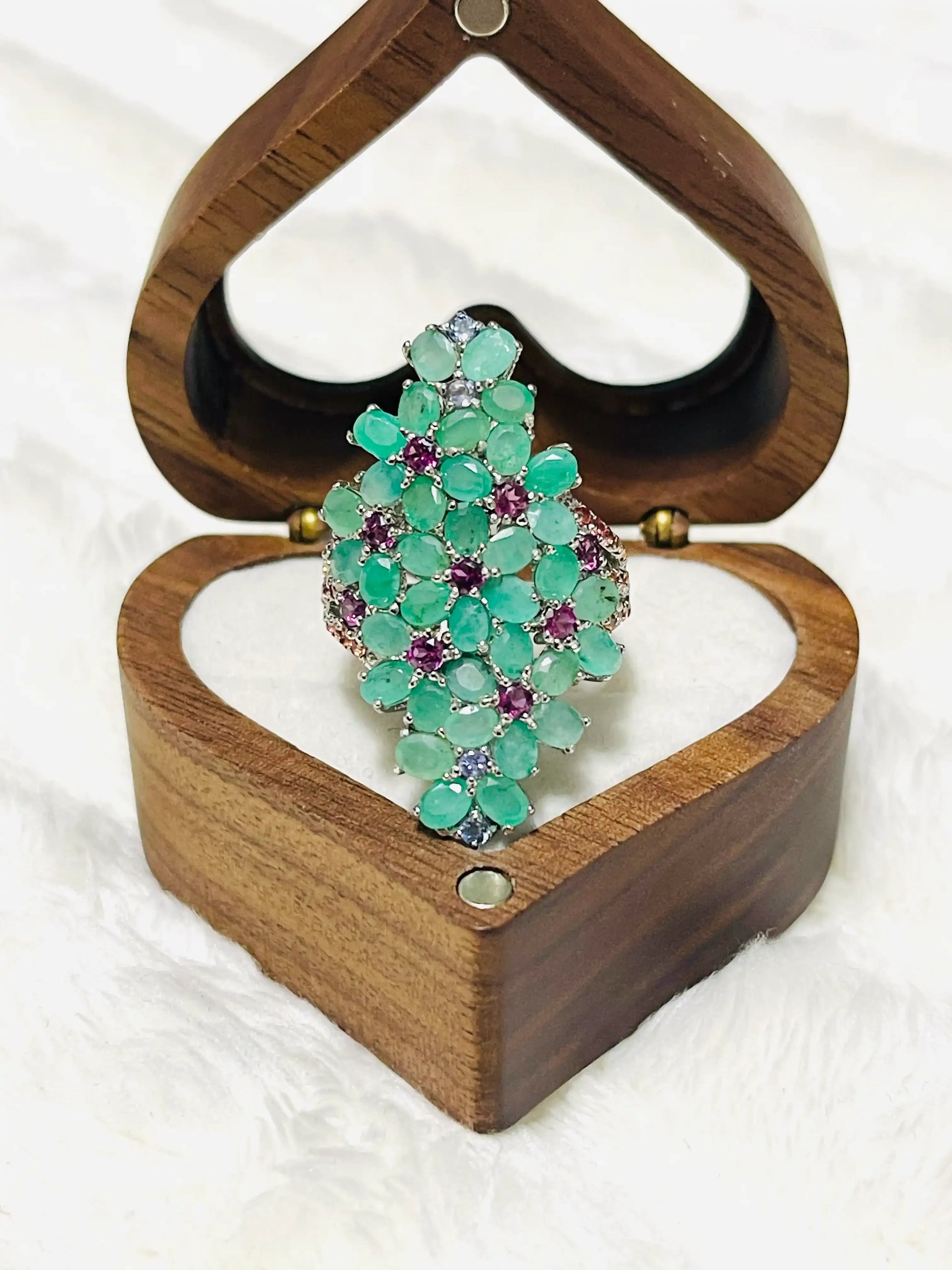Silver ring with a captivating cluster of green emeralds and purple amethyst stones, displayed in a heart-shaped wooden box.