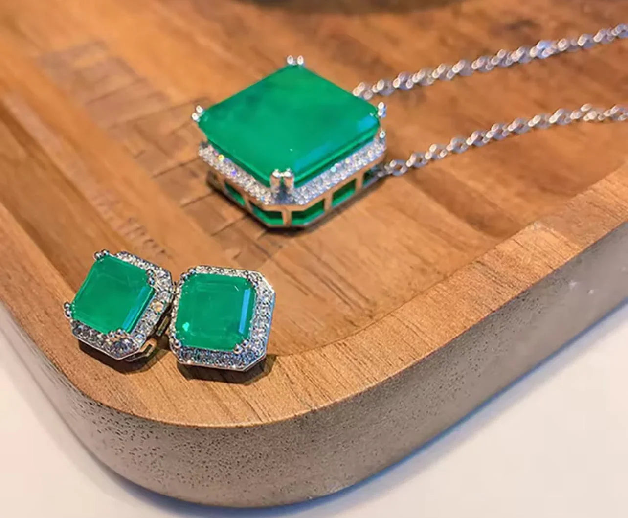 Luxurious emerald and diamond jewelry set, featuring a square pendant and matching earrings, resting on a wooden tray.