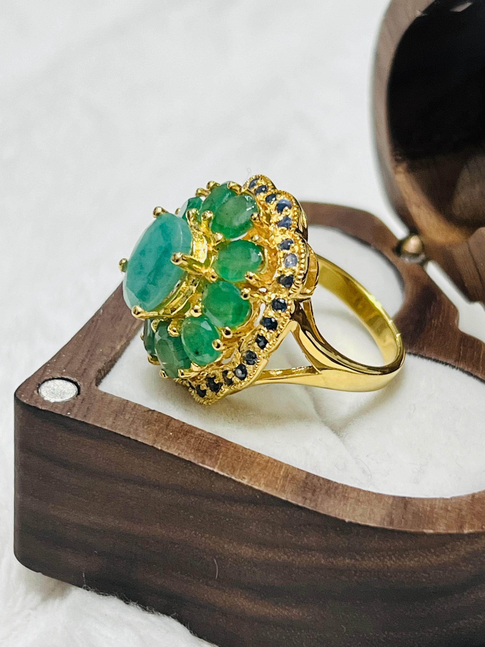 Gold ring with large central emerald, surrounded by smaller emeralds and blue sapphires, displayed in an open wooden jewelry box