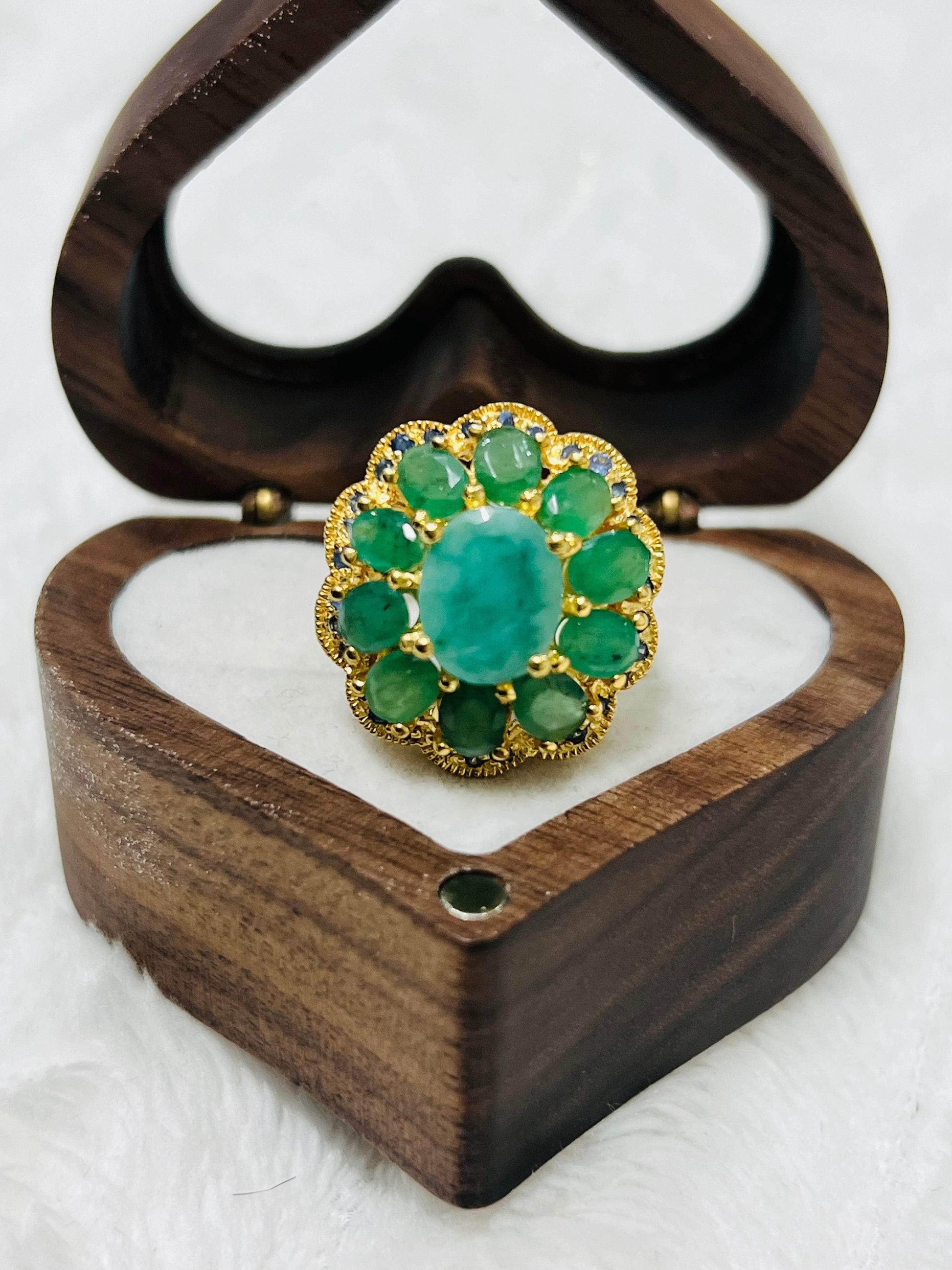 Elegant gold flower ring with emeralds and sapphire details, presented within a charming heart-shaped wooden jewelry box.