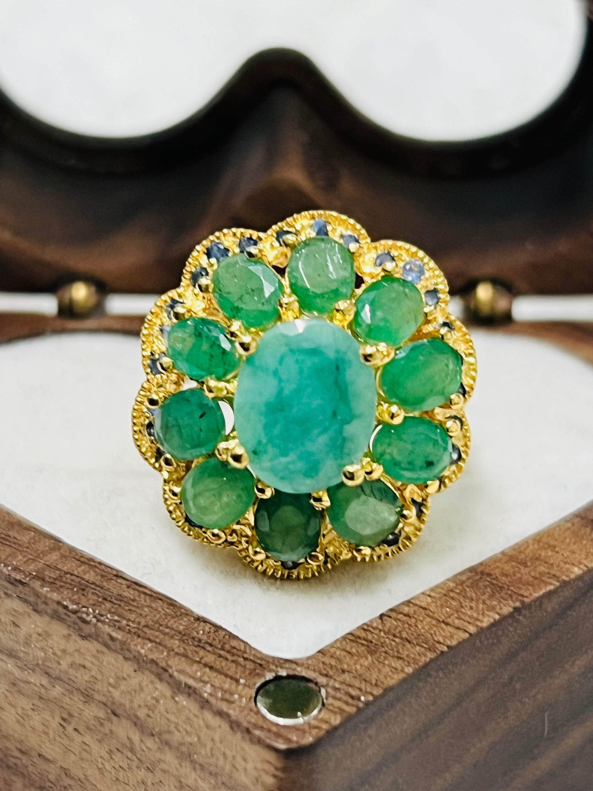 Top-down view of gold cluster ring with central emerald, surrounded by emeralds and blue sapphires, in a wooden jewelry box.