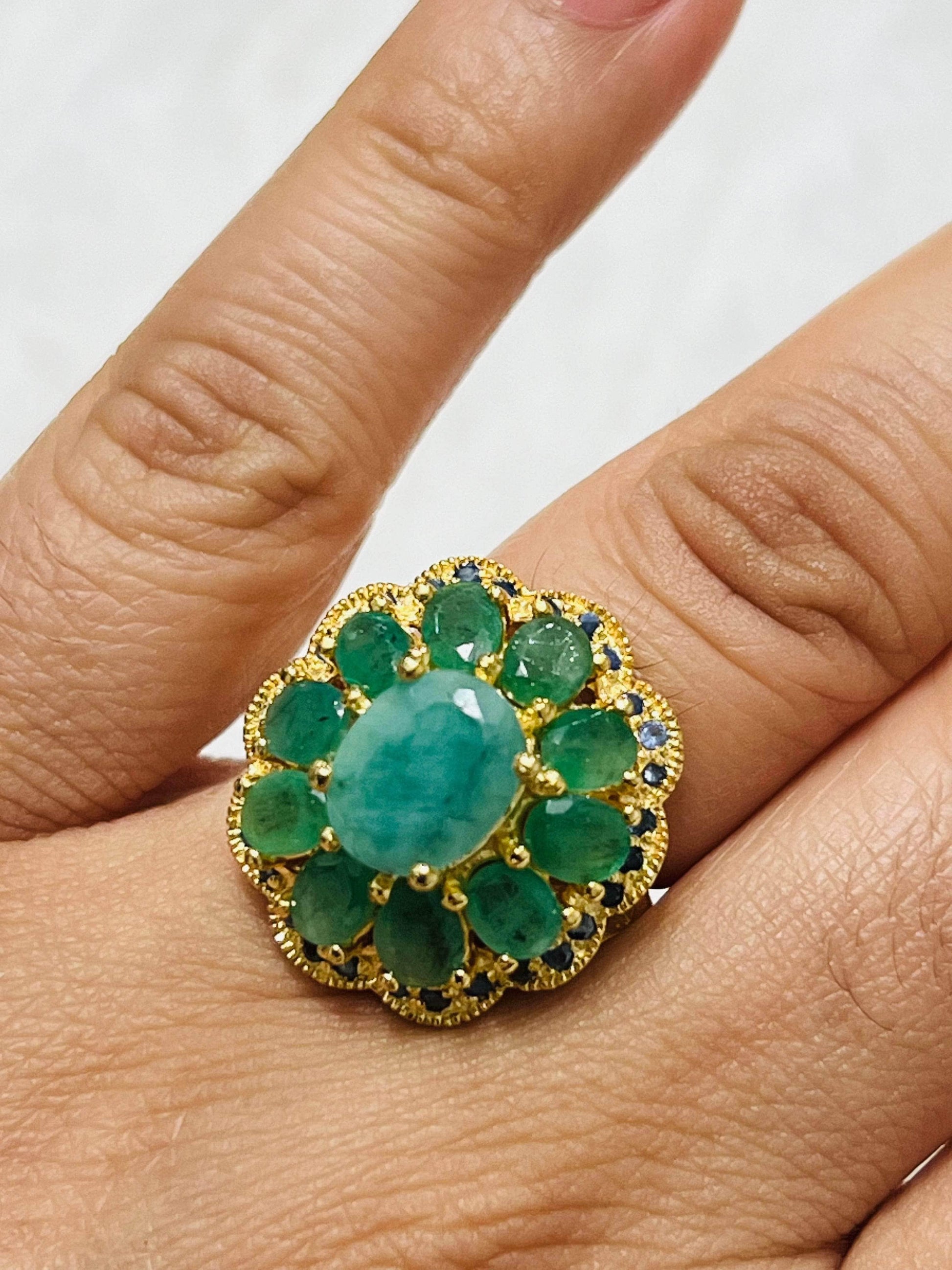 Gold flower ring with a central oval emerald, surrounded by smaller emeralds and blue sapphires, beautifully worn on a finger.