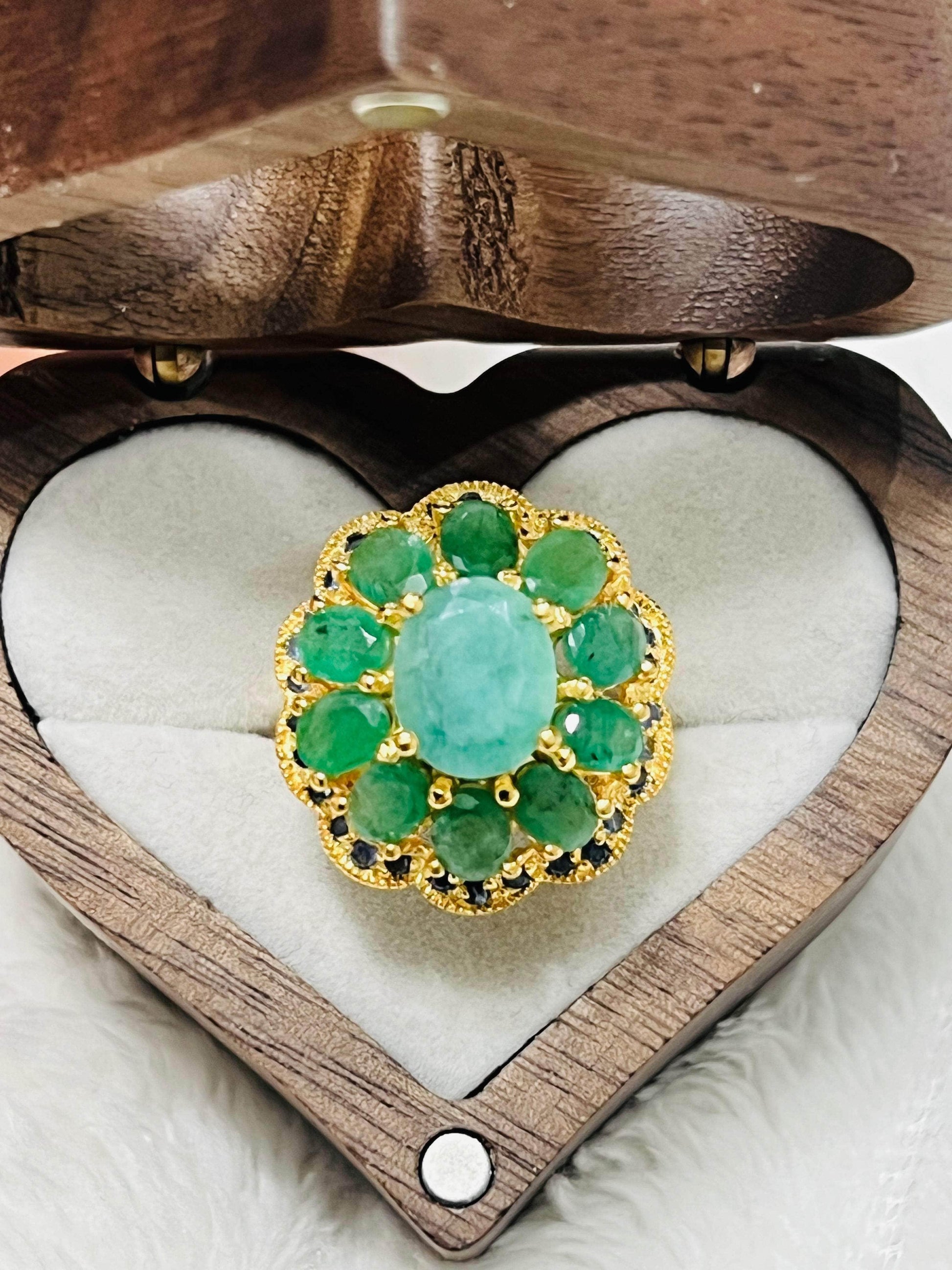 Overhead view of gold ring with central emerald, surrounded by smaller emeralds and blue sapphires in a heart-shaped wooden box