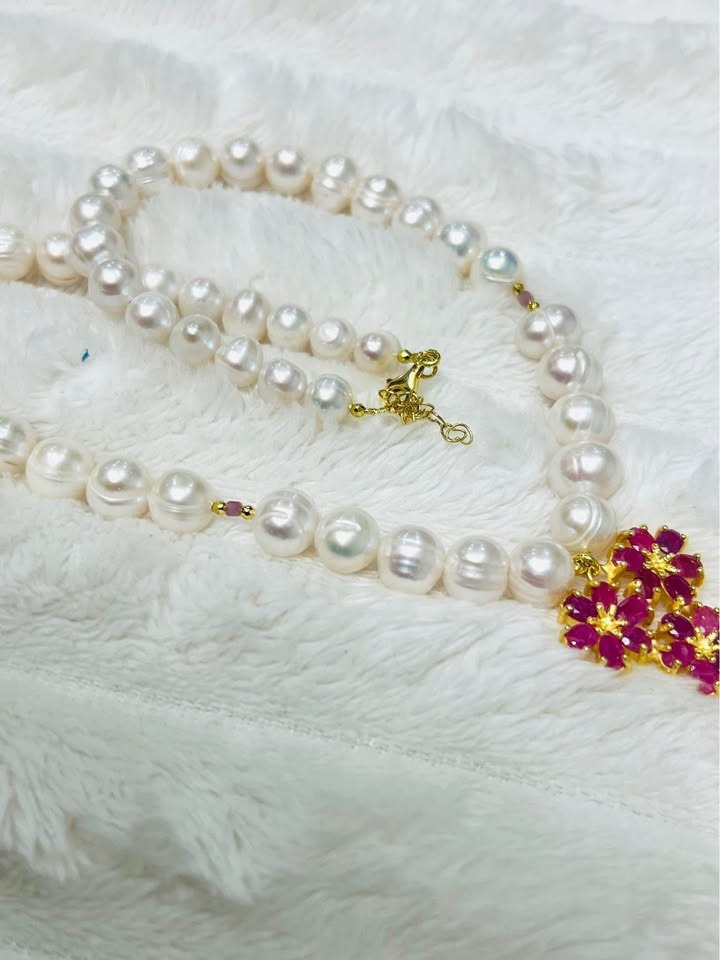 Close-up of natural freshwater pearl and ruby flower necklace with gold clasp on white textured fabric.