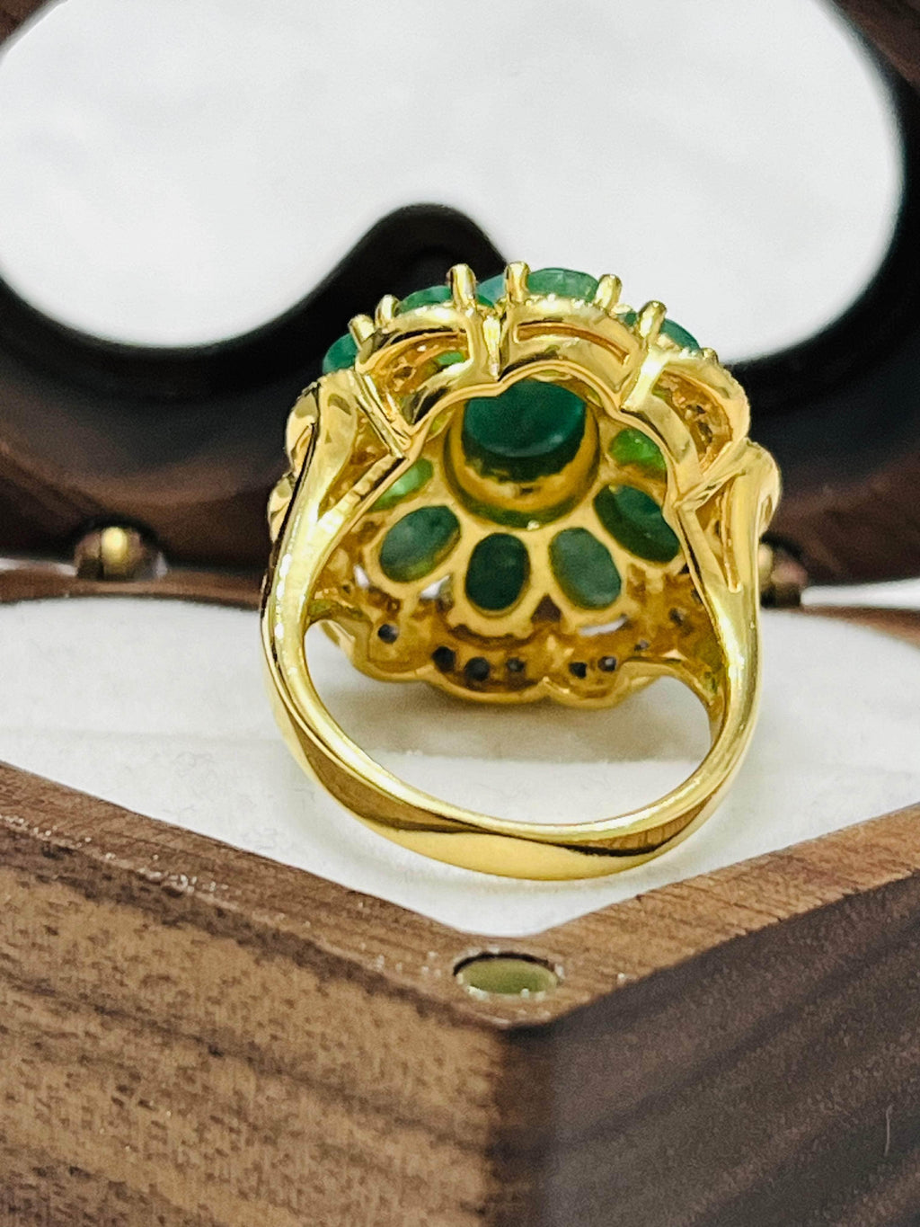 Back view of an intricate gold ring with multiple emeralds and small sapphires, showcasing craftsmanship in a wooden jewelry box.