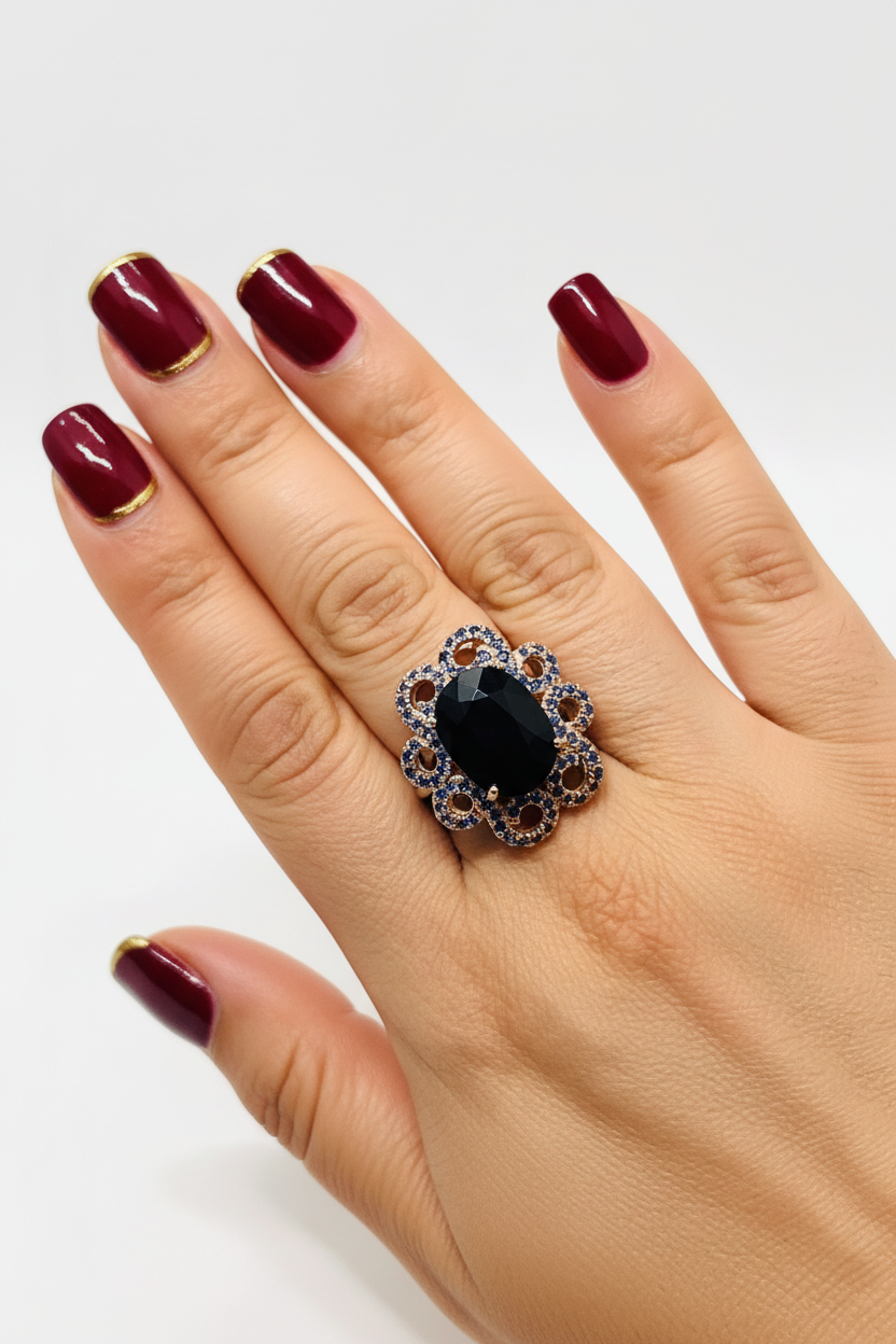 Hand with dark red square nails and gold trim wearing black gemstone ring