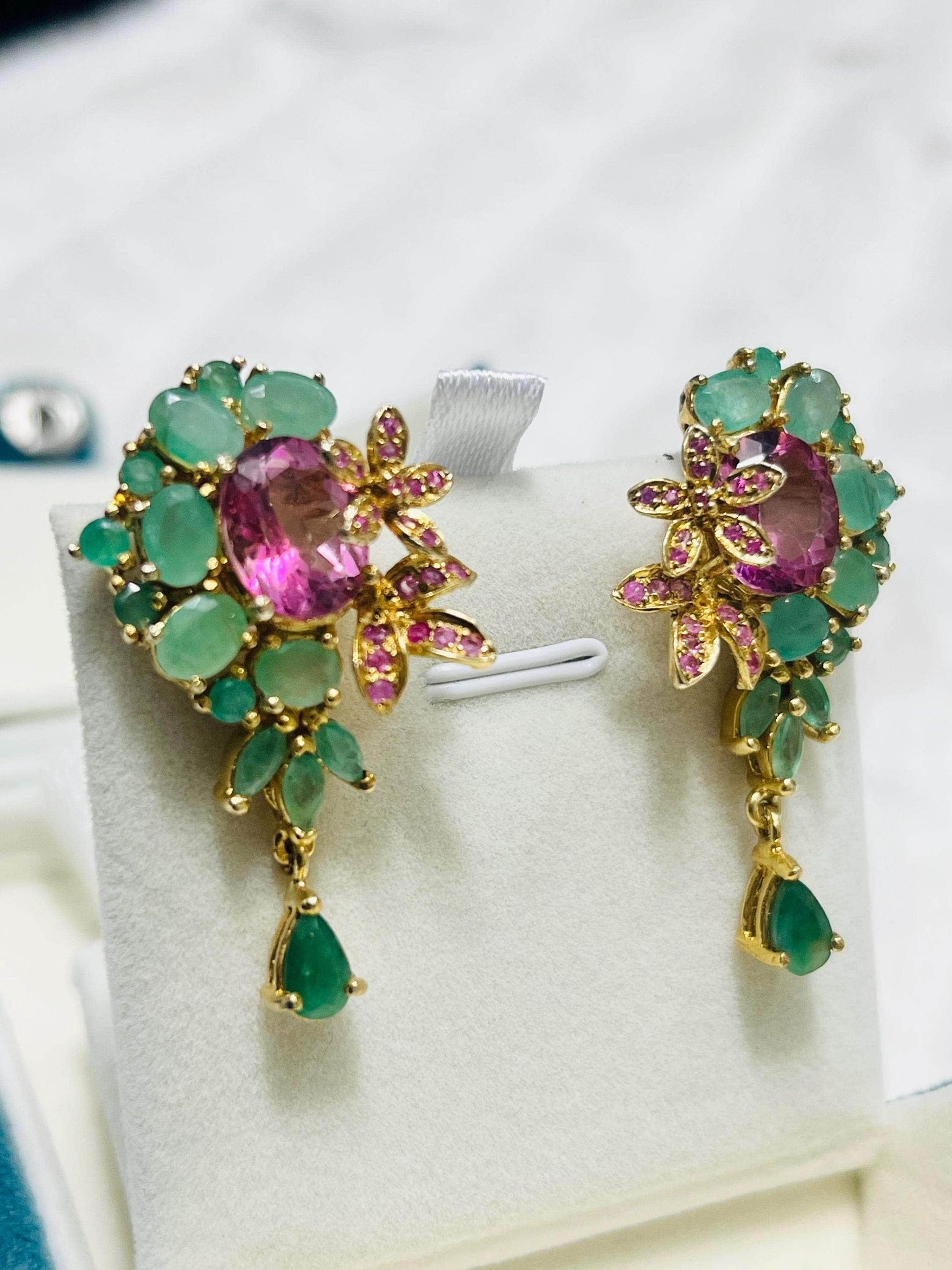 Vibrant Italian earrings featuring green emeralds, pink topaz, and small sapphires on a white display stand.
