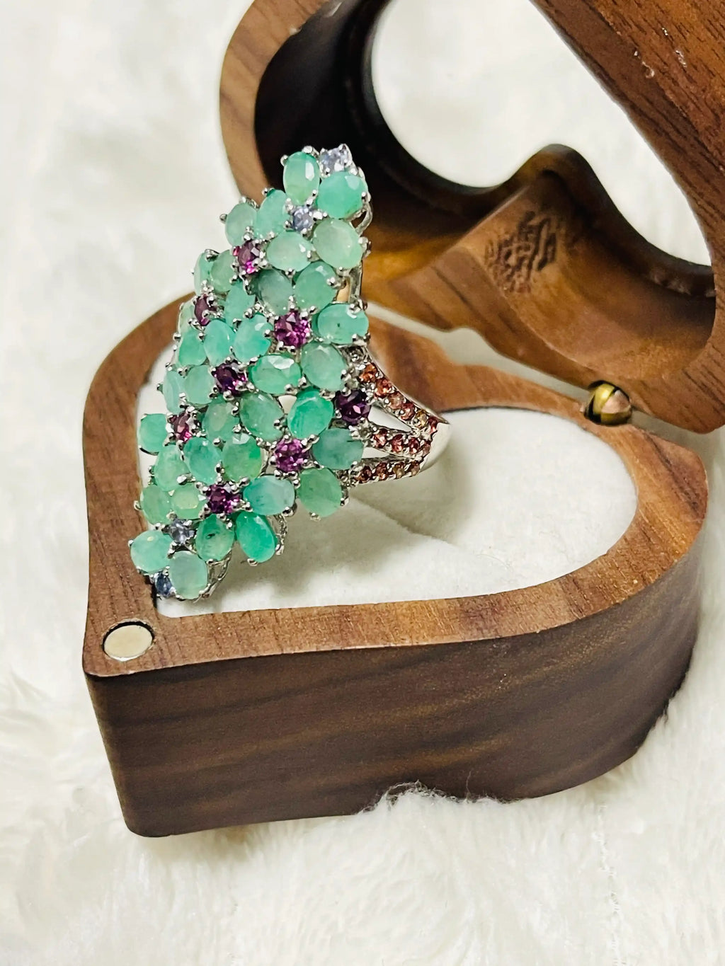 Side view of a large silver cocktail ring with many oval emeralds and purple stones, in a heart-shaped box.