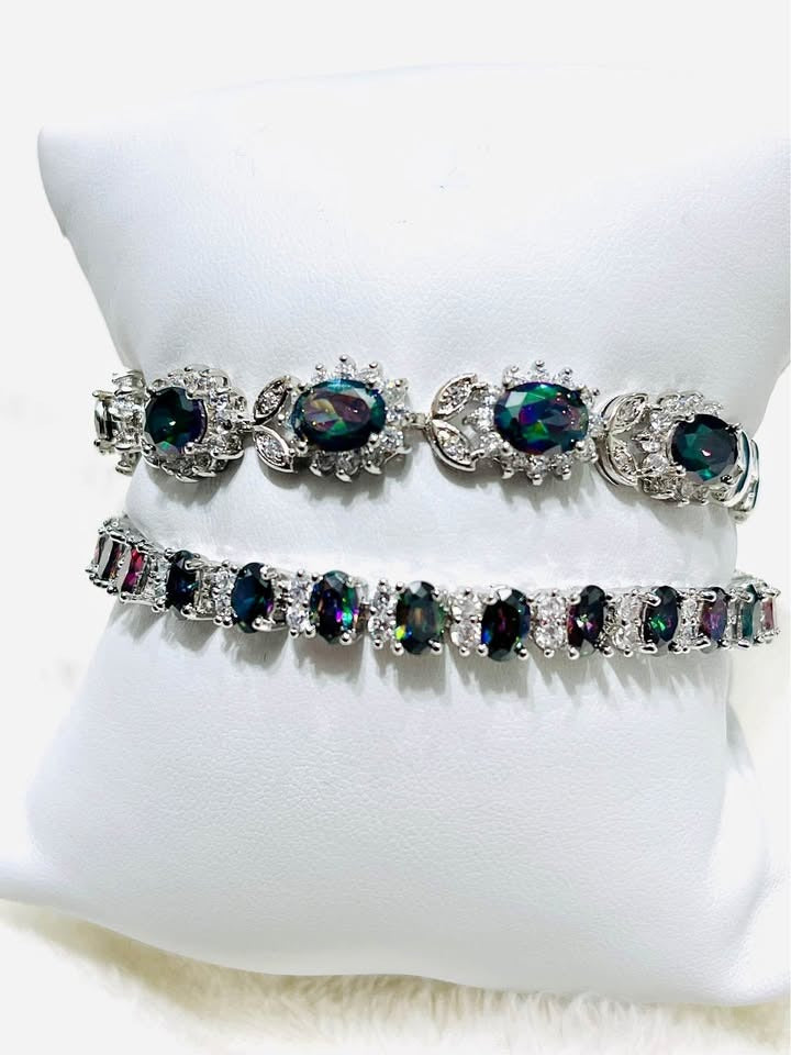  Pair of exquisite mystic topaz sterling silver bracelets, one oval with leaf accents, displayed on a white cushion.