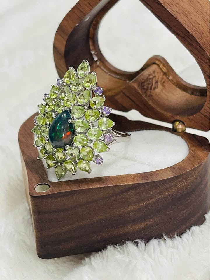 Side view of a peridot and black fire opal ring in a wooden heart-shaped box. Shows the ring's profile.