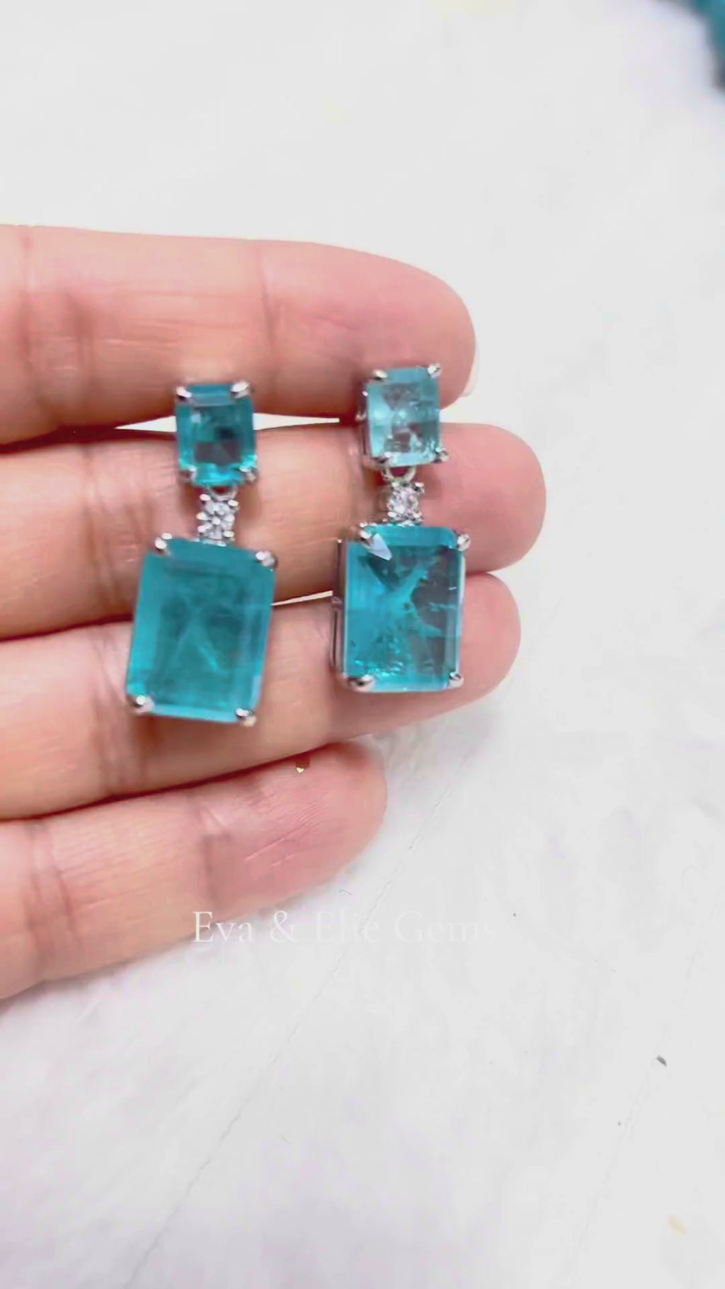 A close-up video showcasing a pair of exquisite drop earrings featuring two large, Paraiba Tourmaline gemstones, with a smaller rectangular stone above, sparkling against a light background.