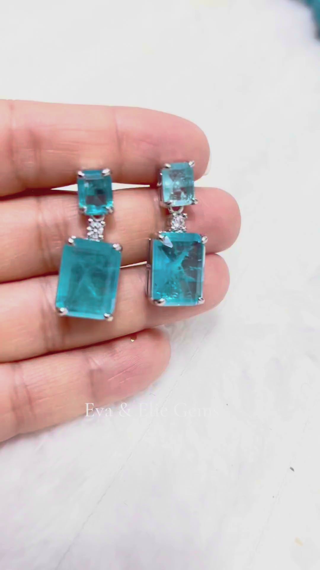 A close-up video showcasing a pair of exquisite drop earrings featuring two large, Paraiba Tourmaline gemstones, with a smaller rectangular stone above, sparkling against a light background.