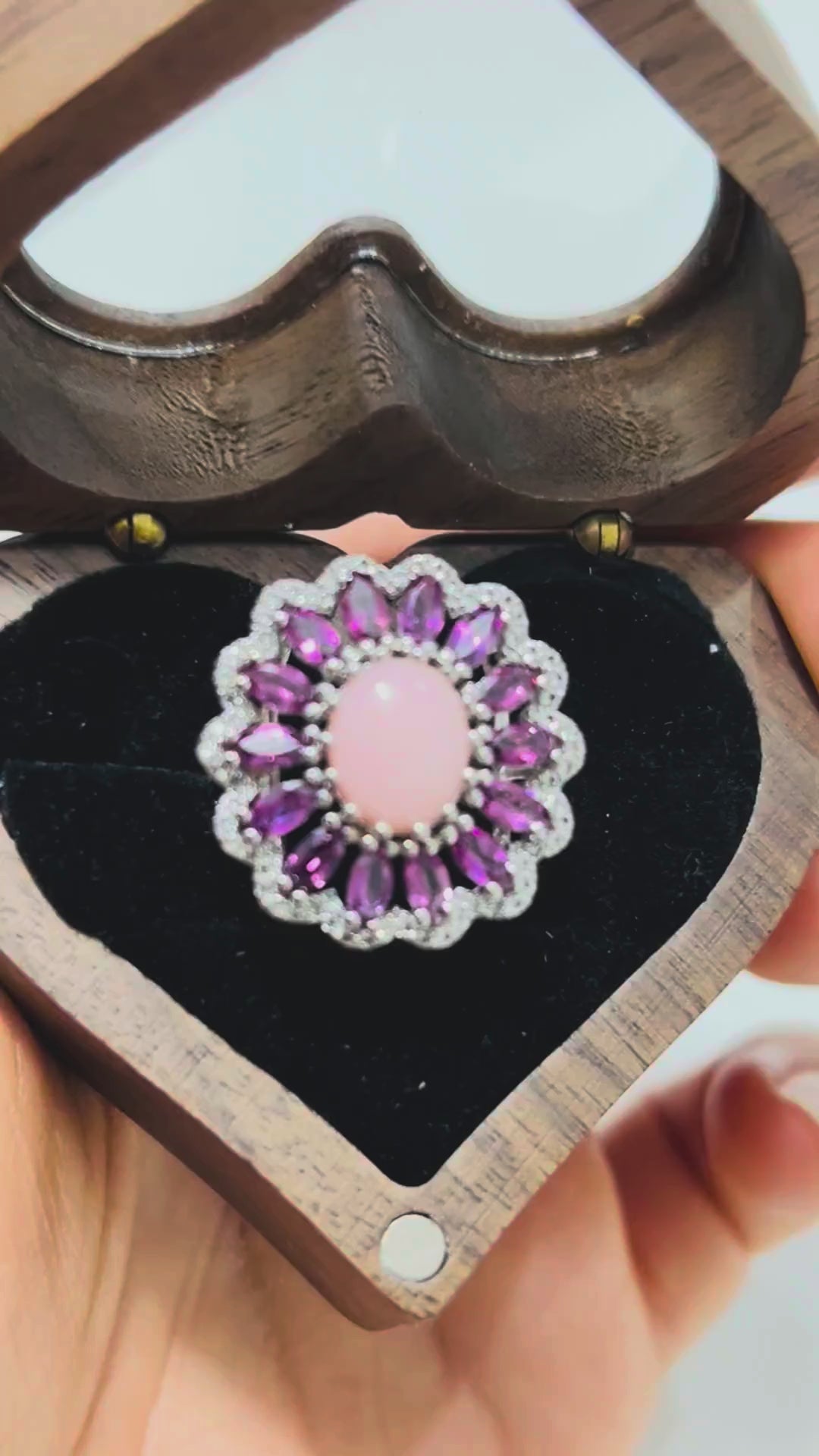 A video showcasing a stunning pink opal and rhodolite garnet ring with a floral halo design, elegantly displayed within a heart-shaped wooden jewelry box.