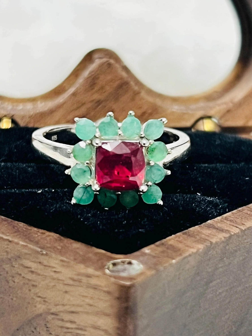Frontal view of a stunning handmade ruby and emerald ring with a floral halo, displayed in a wooden box.