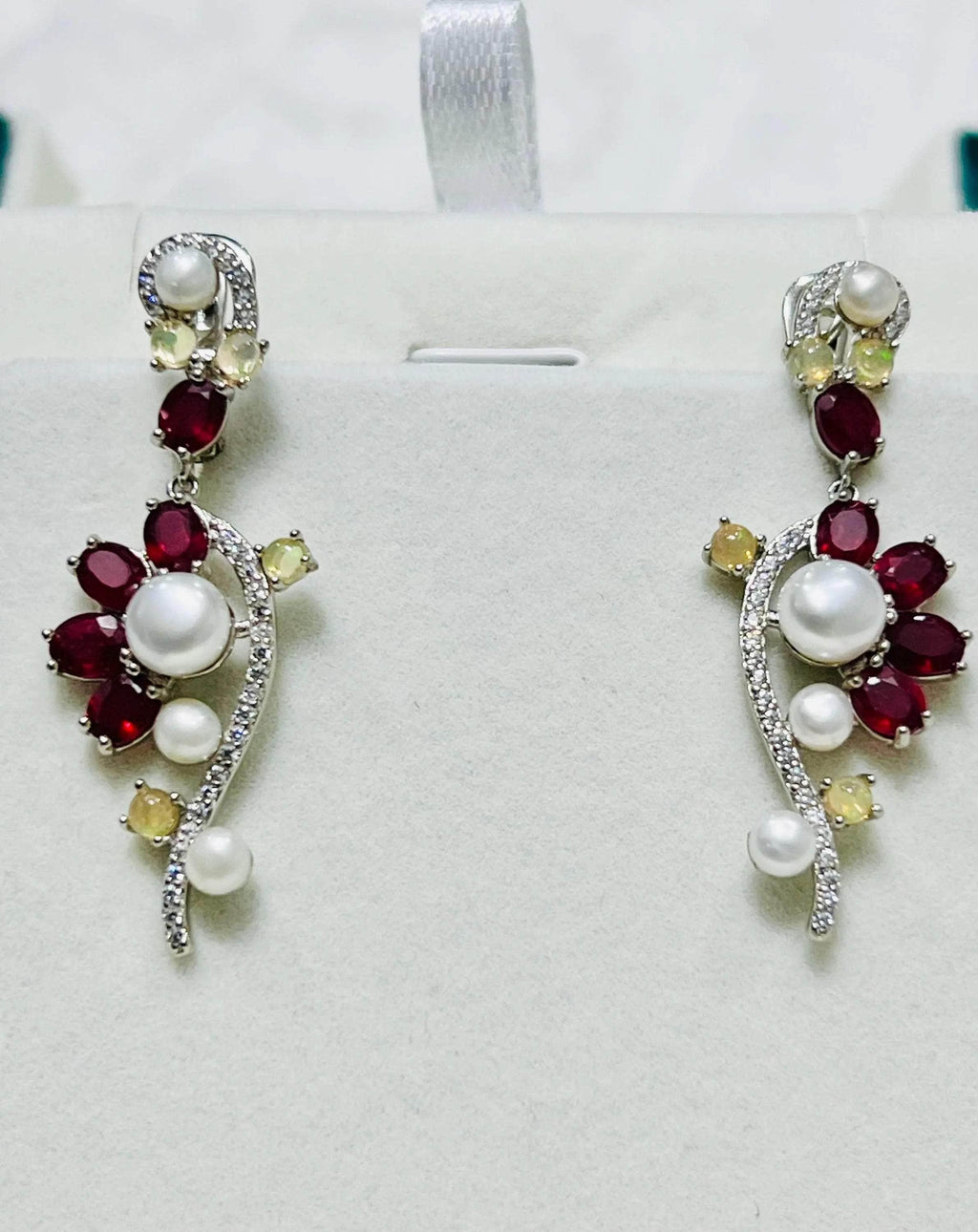 Pair of elegant long earrings with rubies, opals, and pearls, displayed upright on a white jewelry stand.
